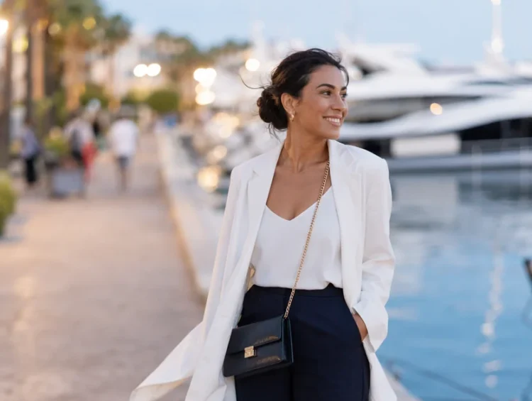 Woman in white blazer and navy trousers walking along a waterfront promenade at dusk with yachts in the background.