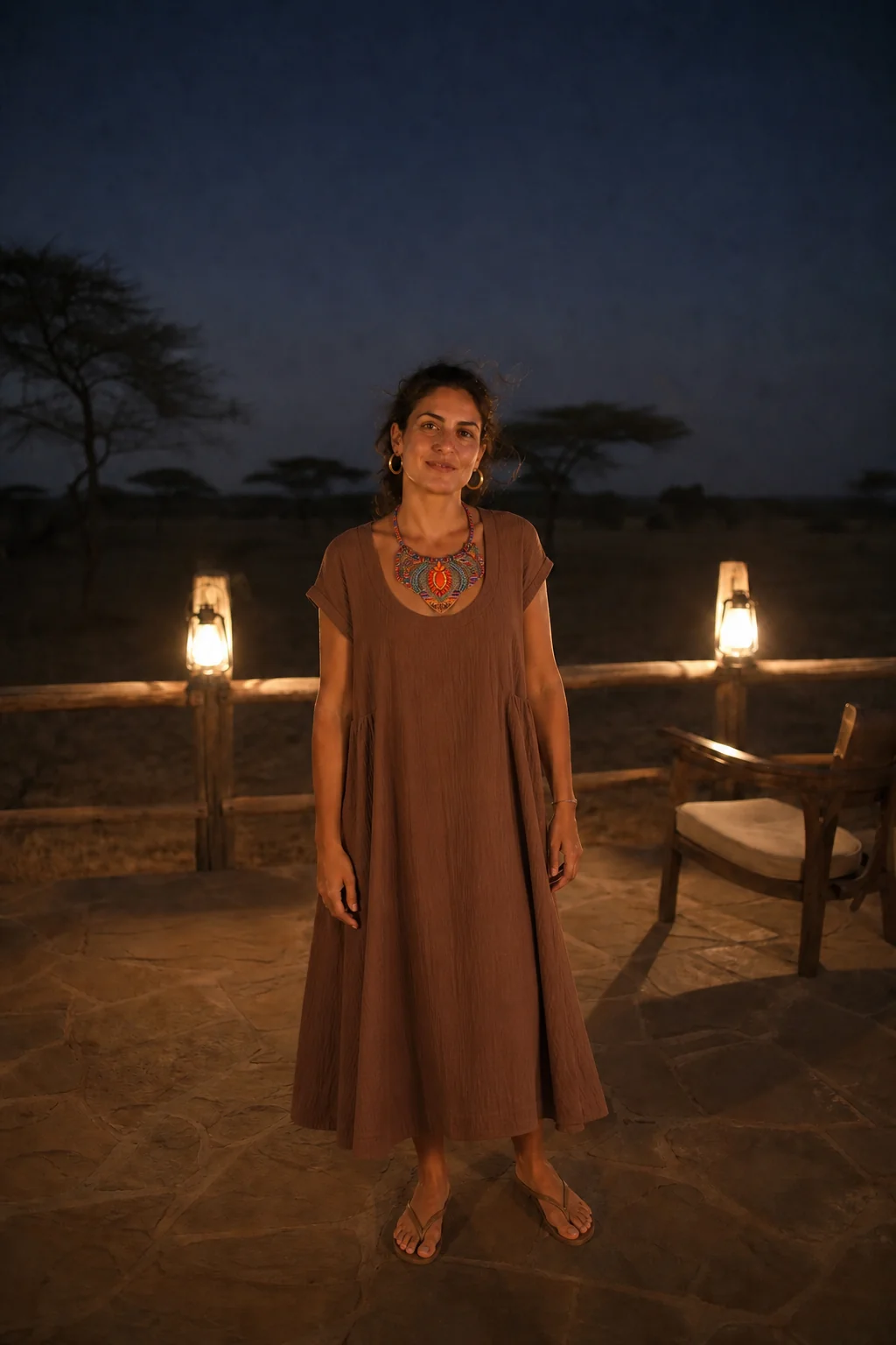 A woman in a brown dress stands on a stone terrace at night, lit by lanterns, with acacia trees in the background.
