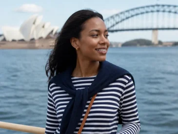 Woman in striped shirt and white shorts standing on ferry railing with Sydney Opera House and Harbour Bridge in background.