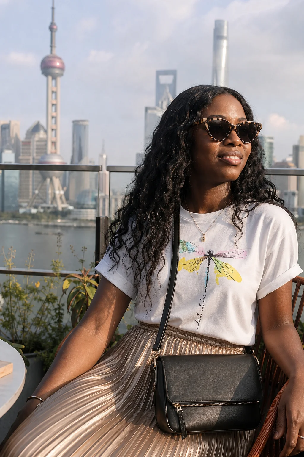 Woman wearing a white graphic tee and pleated gold skirt, sitting on a rooftop with Shanghai skyline in background.