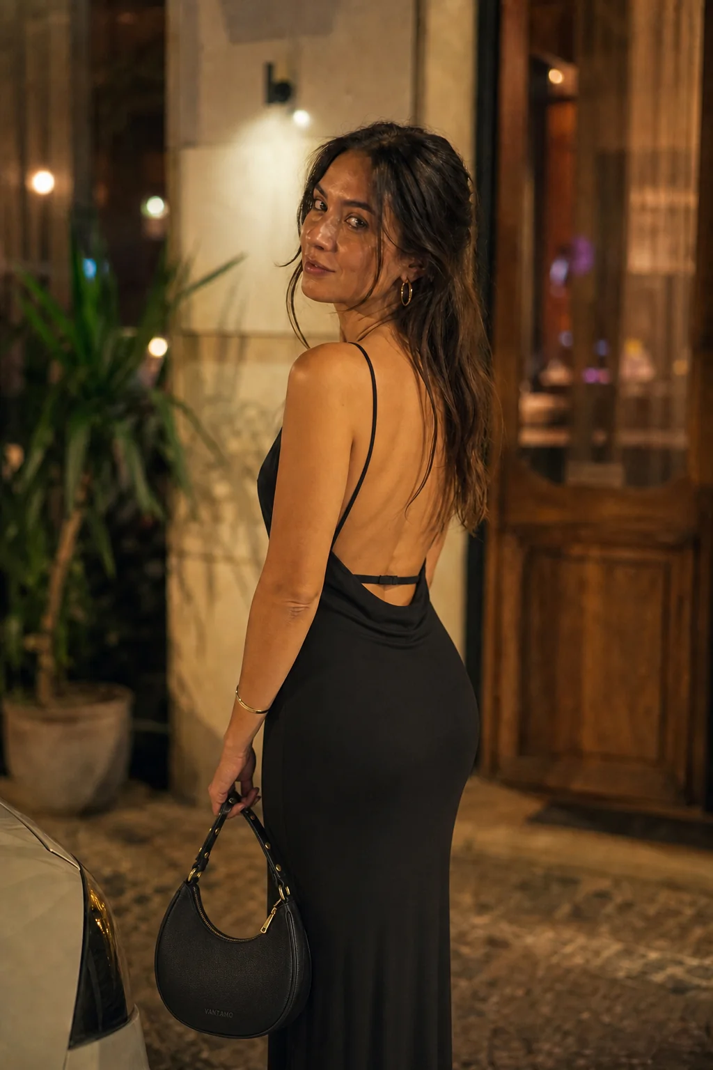 A woman in a black backless dress stands outside at night, holding a small black handbag.