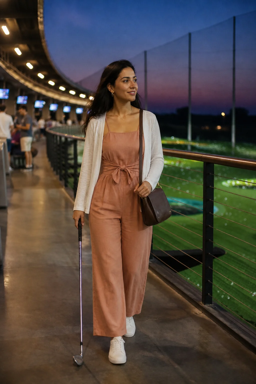 Woman in a peach linen jumpsuit and white cardigan holding a golf club at a Topgolf venue during sunset.